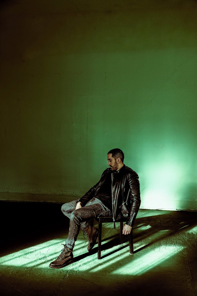 ALP_TD_9631 Cinematic vertical portrait of a man in a black leather jacket sitting on a dark chair in a minimalist studio. A heavy, industrial fluorescent green light flare illuminates the concrete wall behind him, casting long, geometric chair shadows across the floor. The composition uses an inverted color grade with cool highlights and deep, tobacco-toned shadow negative space.