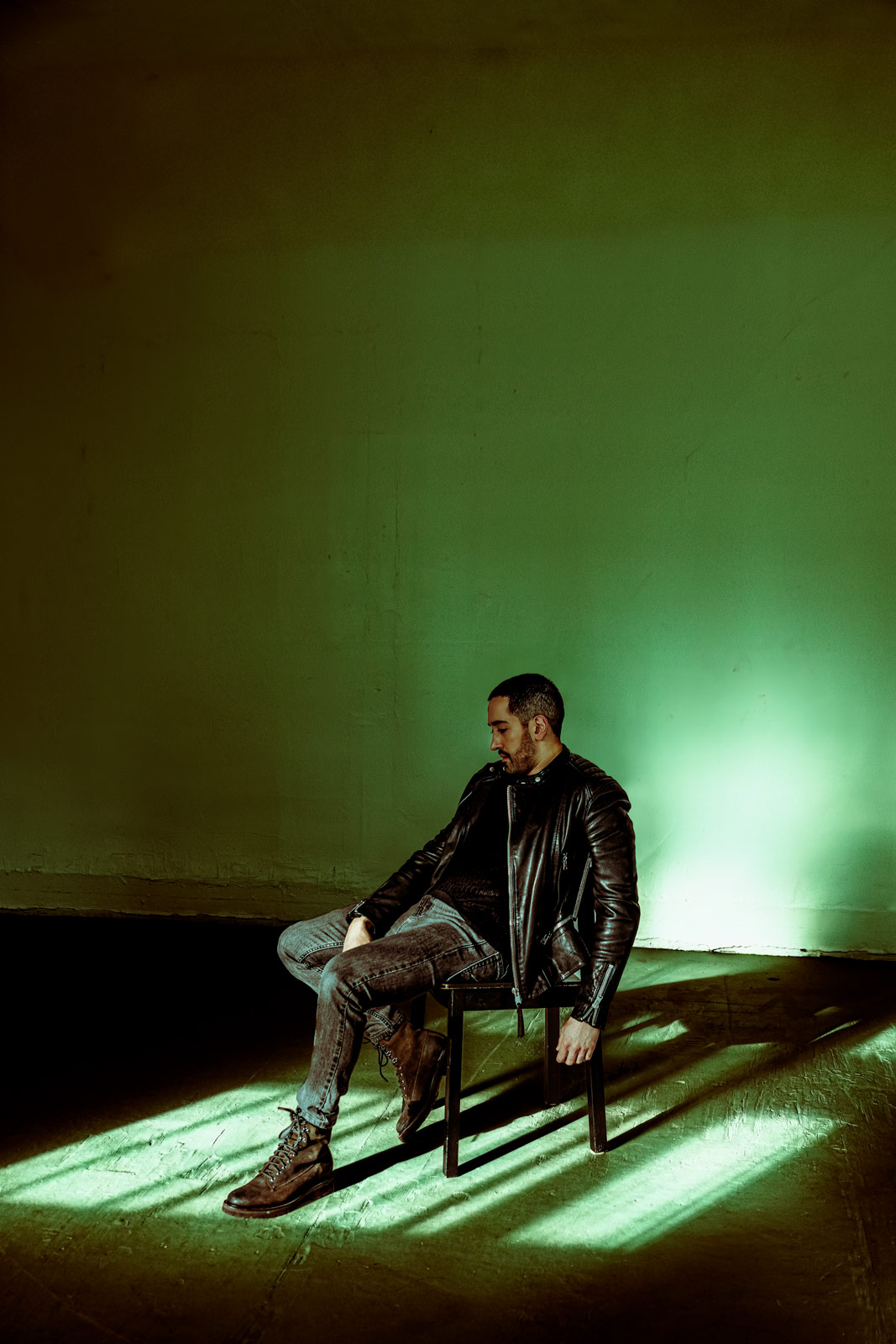 Cinematic vertical portrait of a man in a black leather jacket sitting on a dark chair in a minimalist studio. A heavy, industrial fluorescent green light flare illuminates the concrete wall behind him, casting long, geometric chair shadows across the floor. The composition uses an inverted color grade with cool highlights and deep, tobacco-toned shadow negative space.